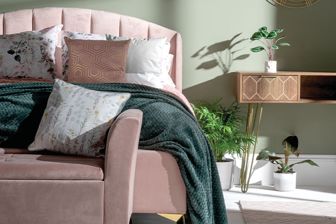 A pink velvet double bed with a matching ottoman at the foot end to the left of the image, with a console table to the right of the image adorned with house plants