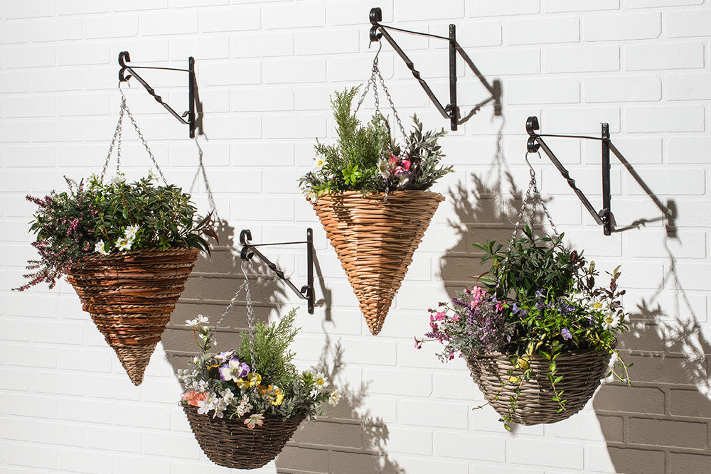 Four hanging baskets filled with vibrant greenery and blooming flowers, suspended from hooks against a white brick wall