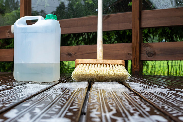 A person using a broom to clean a wooden decking surface, sweeping away debris and dirt, and creating soapy water